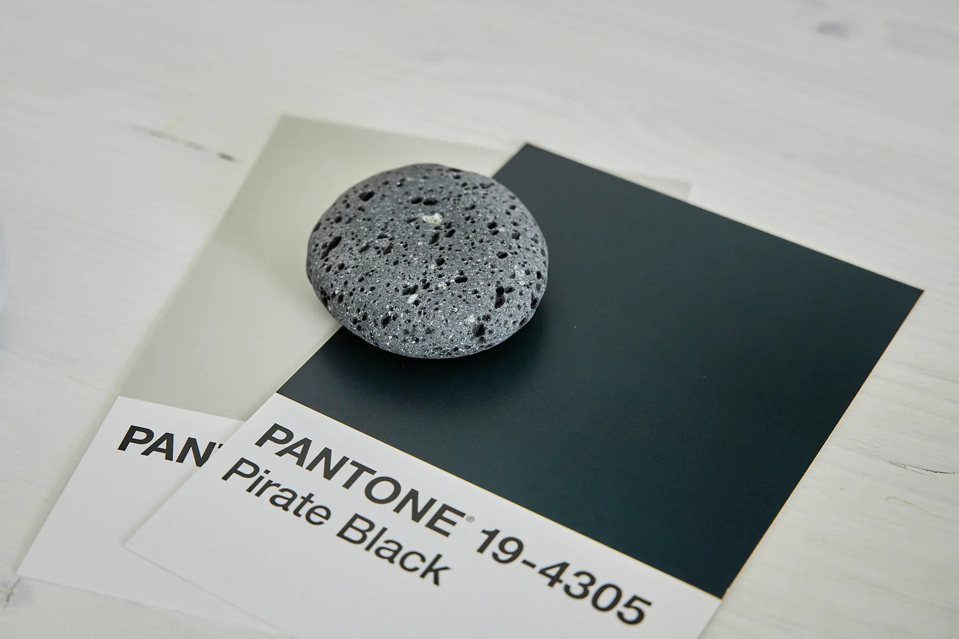 2024-08-18 Nadine Cardenäo 043 Pantone Farbkarte mit den Farben Pirate Black und einem grauen Stein auf einem Tisch.