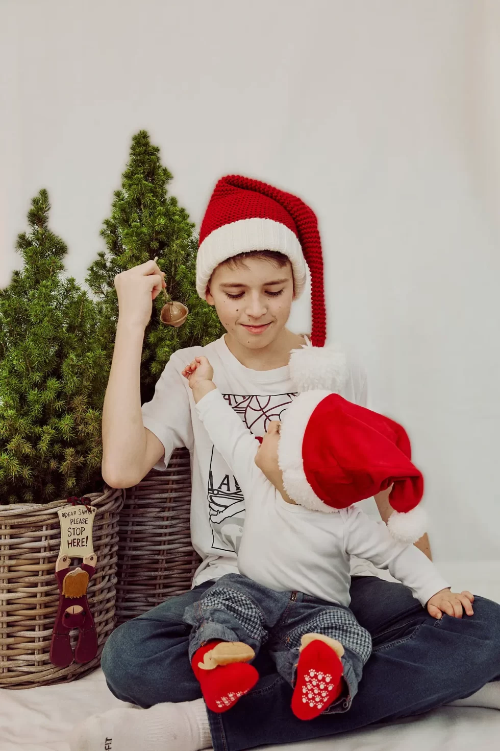 Ein Kind mit einer roten Weihnachtsmütze sitzt auf dem Boden und hält eine Glocke, während ein anderes Kind mit einer ähnlichen Mütze spielt.