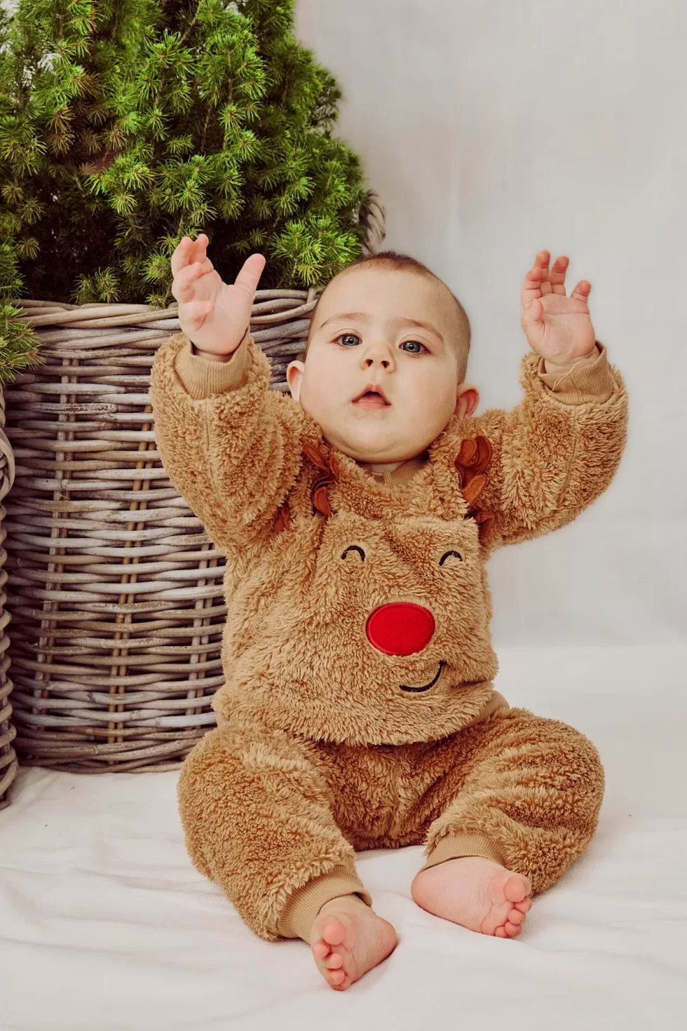 Ein Baby in einem braunen, flauschigen Rentier-Outfit mit roter Nase und ausgestreckten Armen.