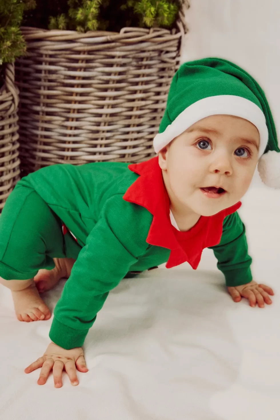 Ein Baby in einem grünen Weihnachtsmannkostüm mit rotem Kragen und einer weißen Mütze sitzt auf dem Boden.