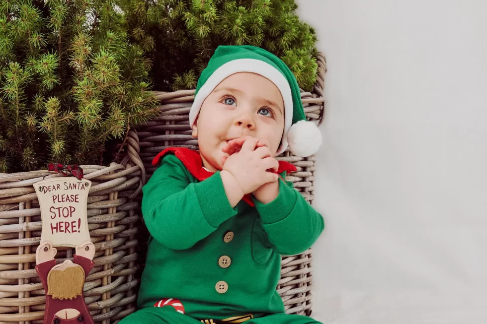 Ein kleines Kind in einem grünen Weihnachtsmannkostüm mit einer roten Schleife und einer Mütze, das an einem Schild mit der Aufschrift 'Dear Santa Please Stop Here' sitzt.