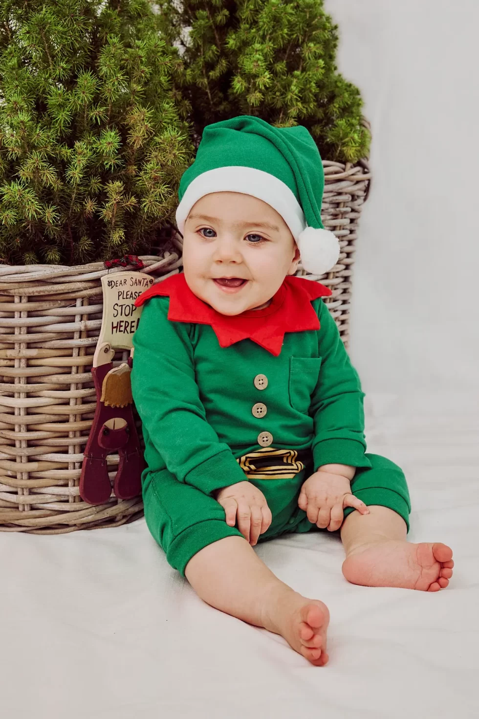 Ein Baby in einem grünen Weihnachtsmannkostüm mit rotem Kragen und einer weißen Mütze sitzt vor einem Korb. Leistungen Austausch