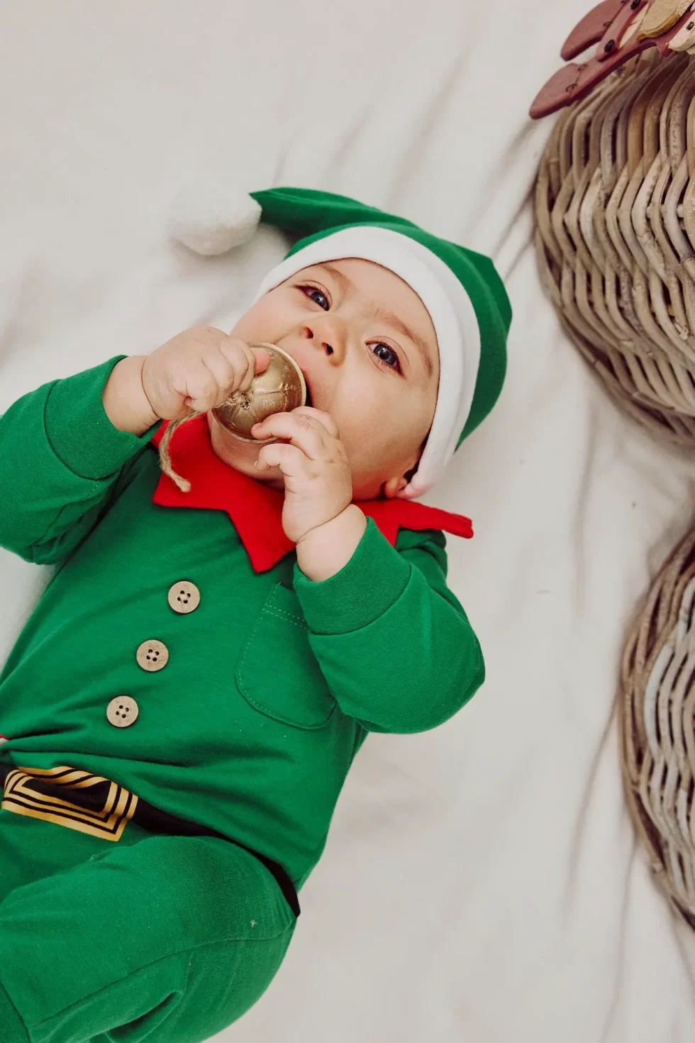 Ein Baby in einem grünen Elfenkostüm mit rotem Kragen hält eine Glocke in der Hand.