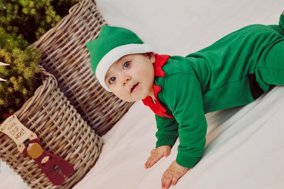 Ein Baby in einem grünen Weihnachtskostüm mit rotem Kragen und einer weißen Mütze auf einem weißen Untergrund.