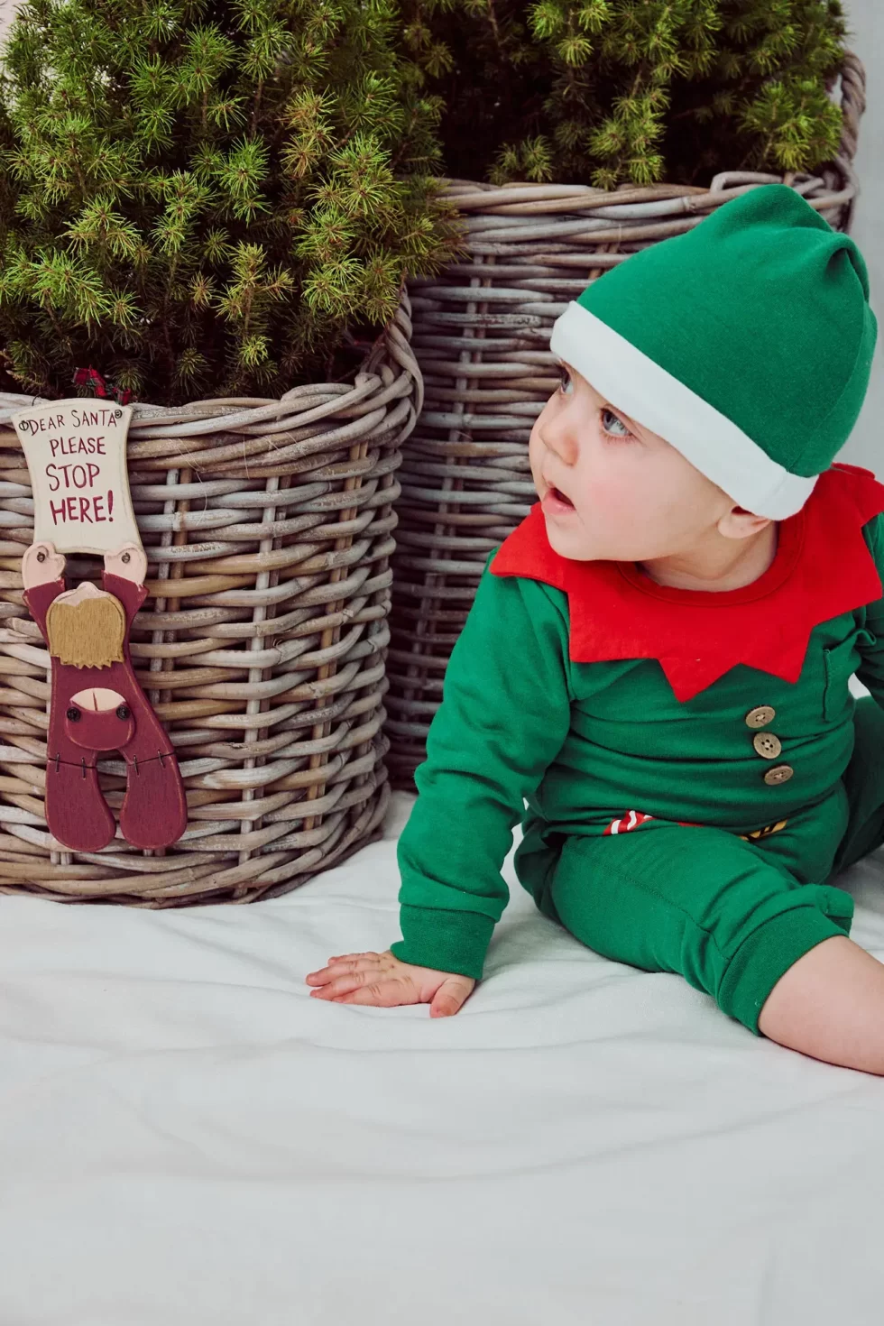 Ein Baby in einem grünen Weihnachtsmannkostüm mit rotem Kragen und Hut sitzt neben einem Korb.